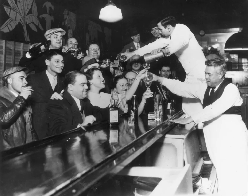 Photo of men and women at the bar in black and white during Prohibition being served alcoholic drinks by 2 bar tenders