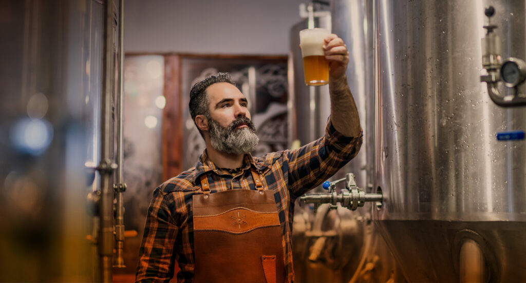 Man standing by brewery tanks holding up and looking at a glass of beer with foam 
