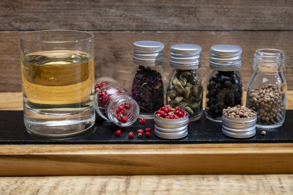 a glass of gin next to various small bottles containing herbs and spices and other ingredients