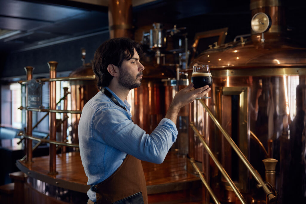 man holding glass full of beer in the production room of a brewery