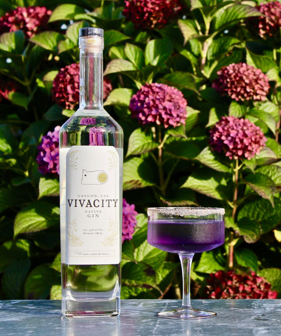 A BOTTLE OF VIVACITY GIN NEXT TO A GLASS HALF FULL IN FRONT OF A FLOWERED BUSH