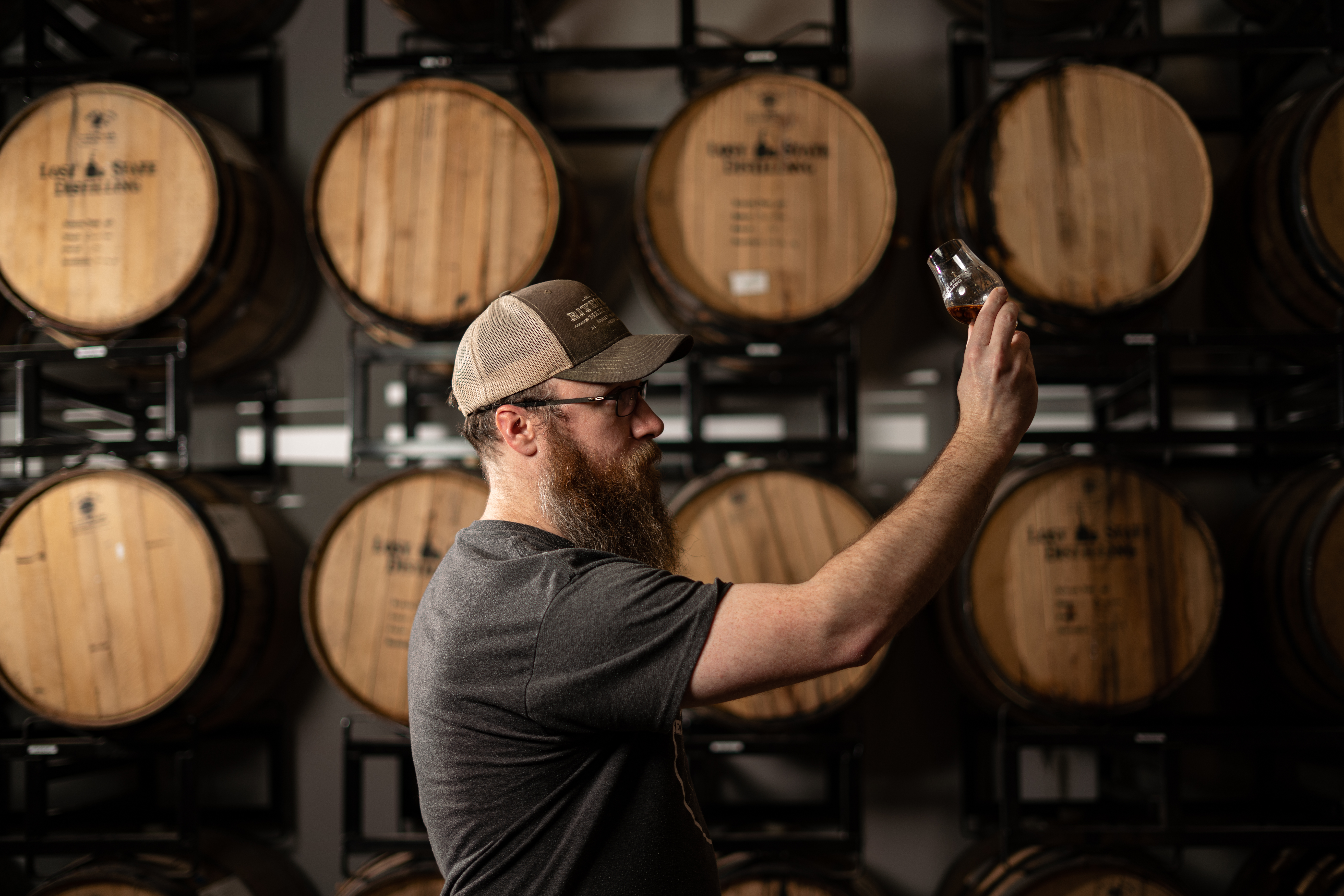 one of the owners at Lost State Distilling inspecting a glass of spirits in front of a rack of barrels