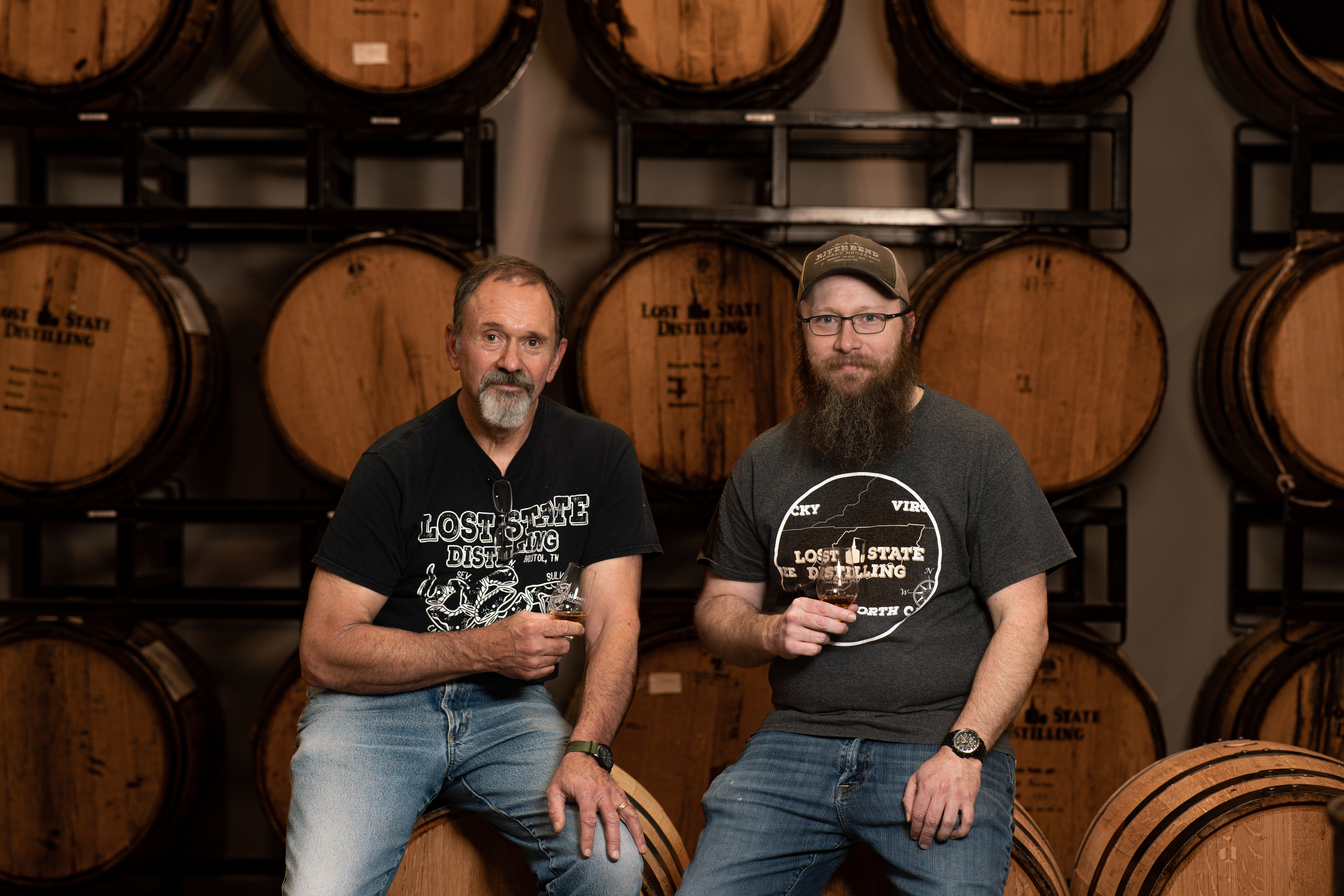 THE OWNERS OF LOST STATE DISTILLING SITTING ON BARRELS WITH A GLASS OF SPIRITS IN THEIR HANDS